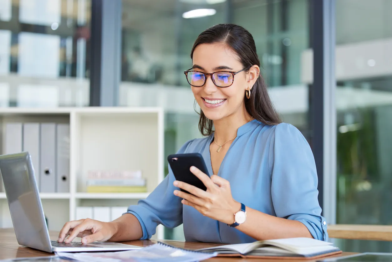 Mujer ejecutiva sentada en su escritorio viendo su celular inteligente, frente a su laptop.