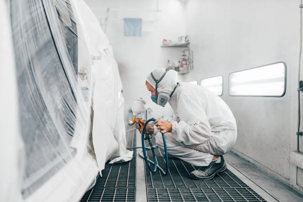Trabajadora en uniforme de protección pintando un coche en una cabina de pintura.