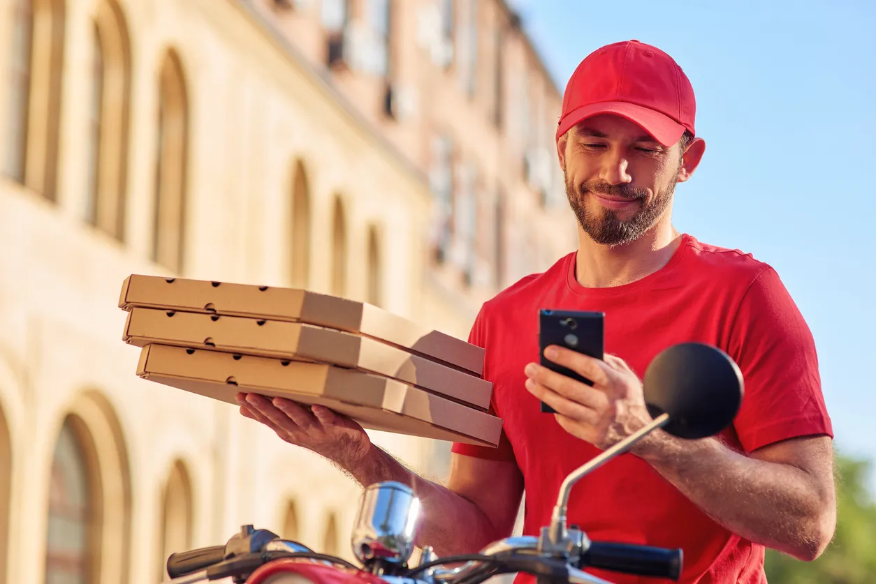 Joven repartidor alegre con uniforme rojo sentado en un scooter y usando un teléfono inteligente de gama media