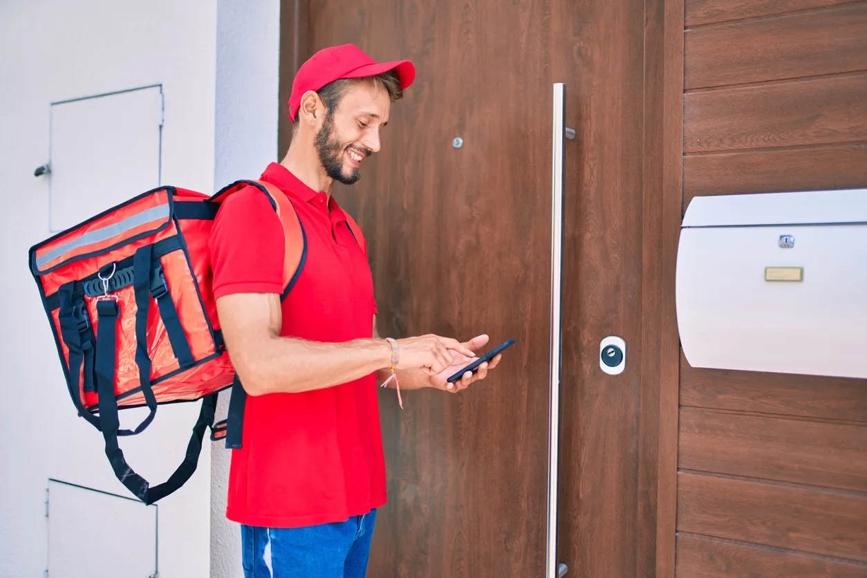 Repartidor caucásico con uniforme rojo y mochila de reparto, sonriendo feliz al aire libre usando un celular sencillo.