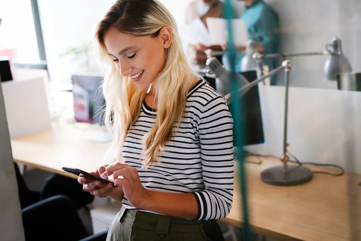 Joven sonriente mirando en un teléfono inteligente durante el trabajo en la oficina