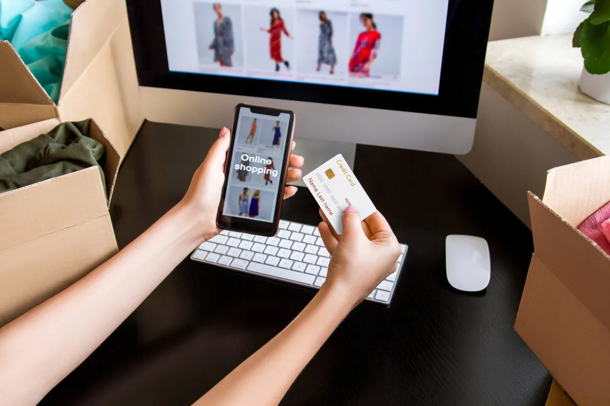 Manos de persona con celular y tarjeta bancaria en manos frente a pantalla de computadora mostrando un tienda online