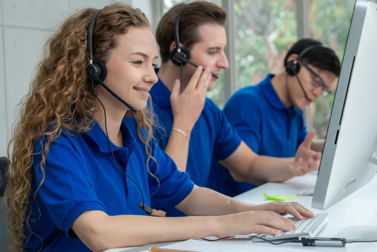 Tres oficinistas con playera tipo polo azul atendiendo llamadas con auriculares y pantalla al frente