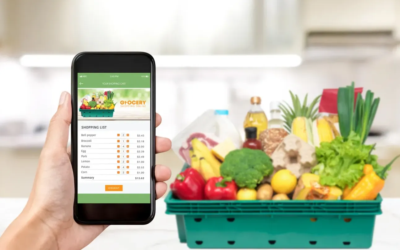 Mano sosteniendo un celular con la pantalla en una tienda en linea de abarrotes. Al fondo fruta diversa en una caja verde.