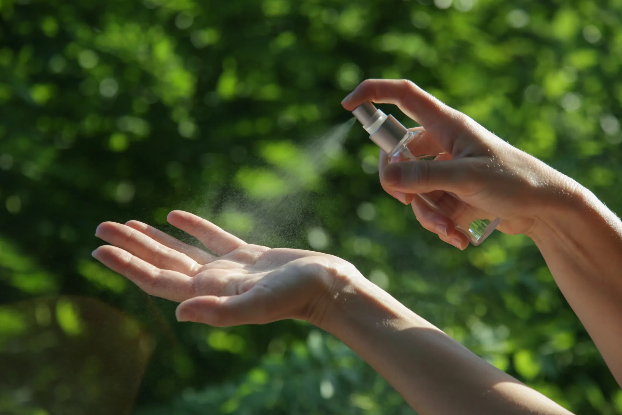 Se muestran una mano aplicandóse repelente contra insectos sobre su otra mano. Al fondo una arbón con gran follaje verde.