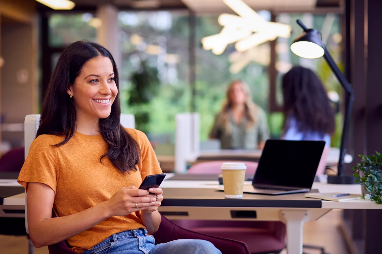 Joven mujer con blusa naranja y pantalón de mezclilla sentada con su celular moderno en ambas manos.