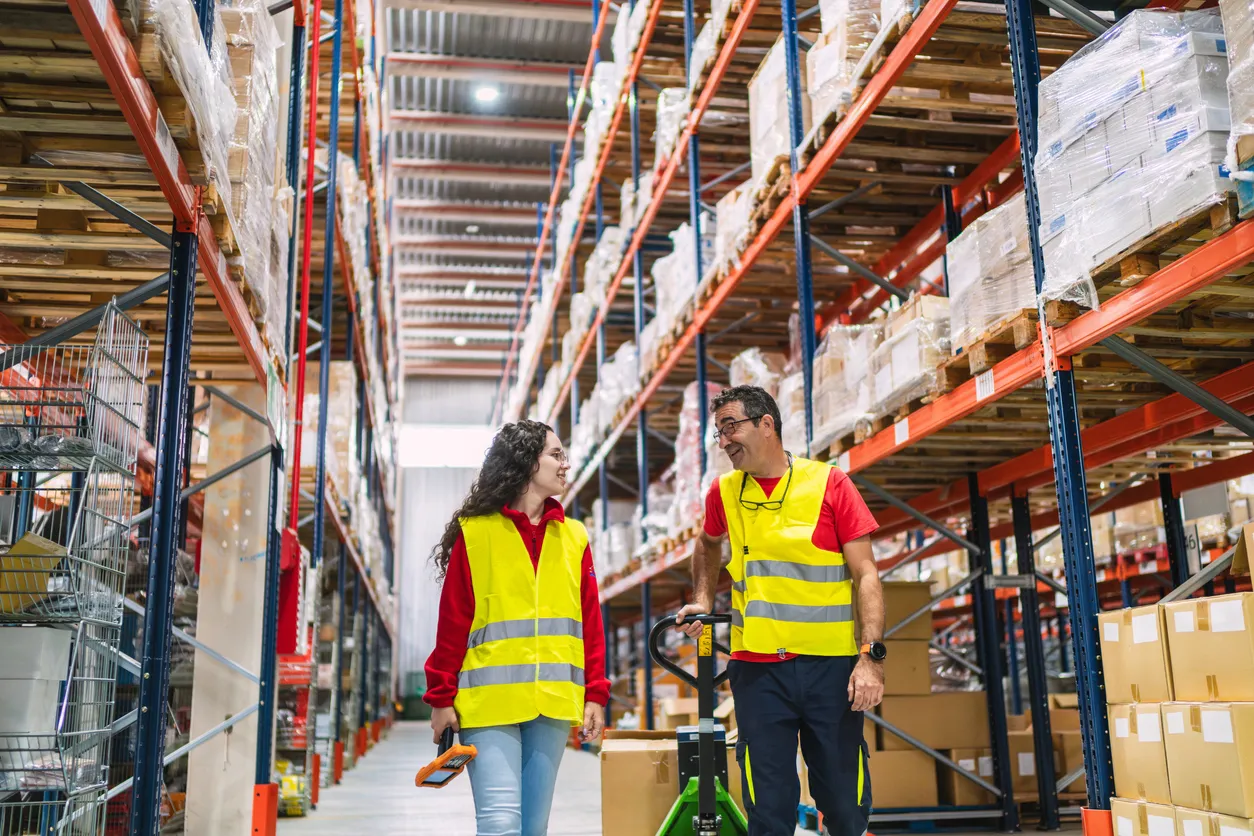 Gran almacen de abarrotes. Dos trabajadores con chaleco de seguridad amarrillo conversan.