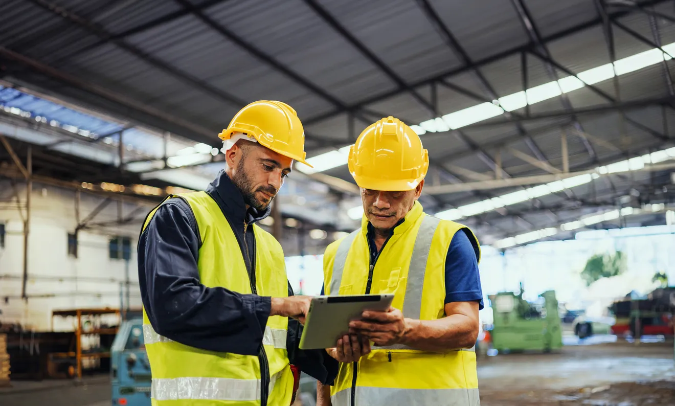 Dos trabajadores con chaleco y casco de seguridad amarillos en fábrica con una gran nave indistrial. Se usan air cooler